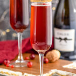 two glasses of Cranberry Pomegranate Champagne Cocktail recipe rimmed with gold sprinkles surrounded by pomegranate seeds and cranberries