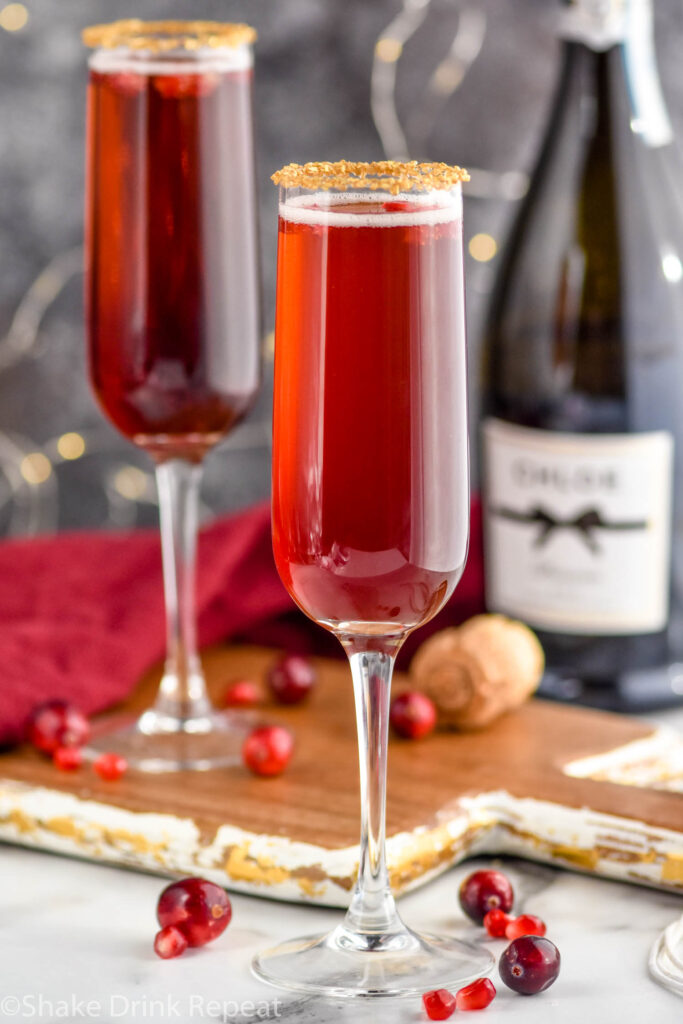 two glasses of Cranberry Pomegranate Champagne Cocktail recipe rimmed with gold sprinkles surrounded by pomegranate seeds and cranberries