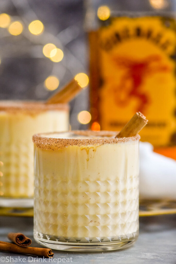 two glasses of Fireball Eggnog with cinnamon sugar rim, cinnamon stick garnish, and bottle of Fireball whiskey in the background