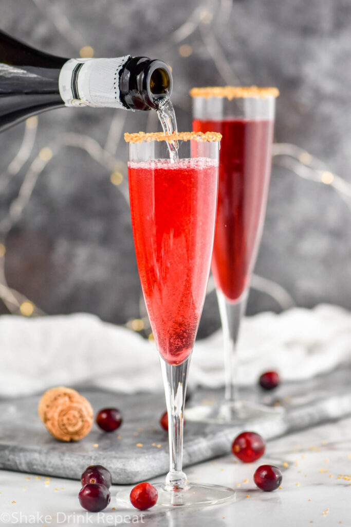 pouring a bottle of Champagne into a glass of Poinsettia Cocktail recipe garnished with gold sprinkles and cranberries