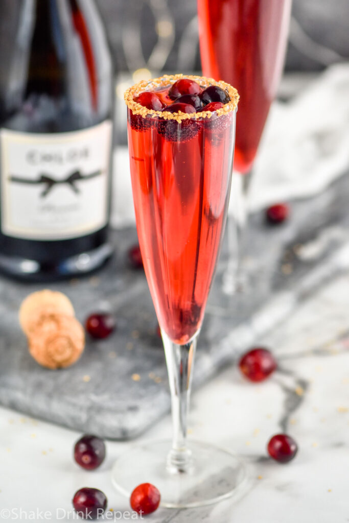 two champagne flutes of Poinsettia Cocktail recipe garnished with gold sprinkles and cranberries with bottle of champagne in the background