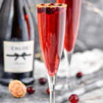 two champagne flutes of Poinsettia Cocktail recipe garnished with gold sprinkles and cranberries with bottle of champagne in the background