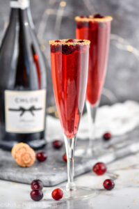 two champagne flutes of Poinsettia Cocktail recipe garnished with gold sprinkles and cranberries with bottle of champagne in the background