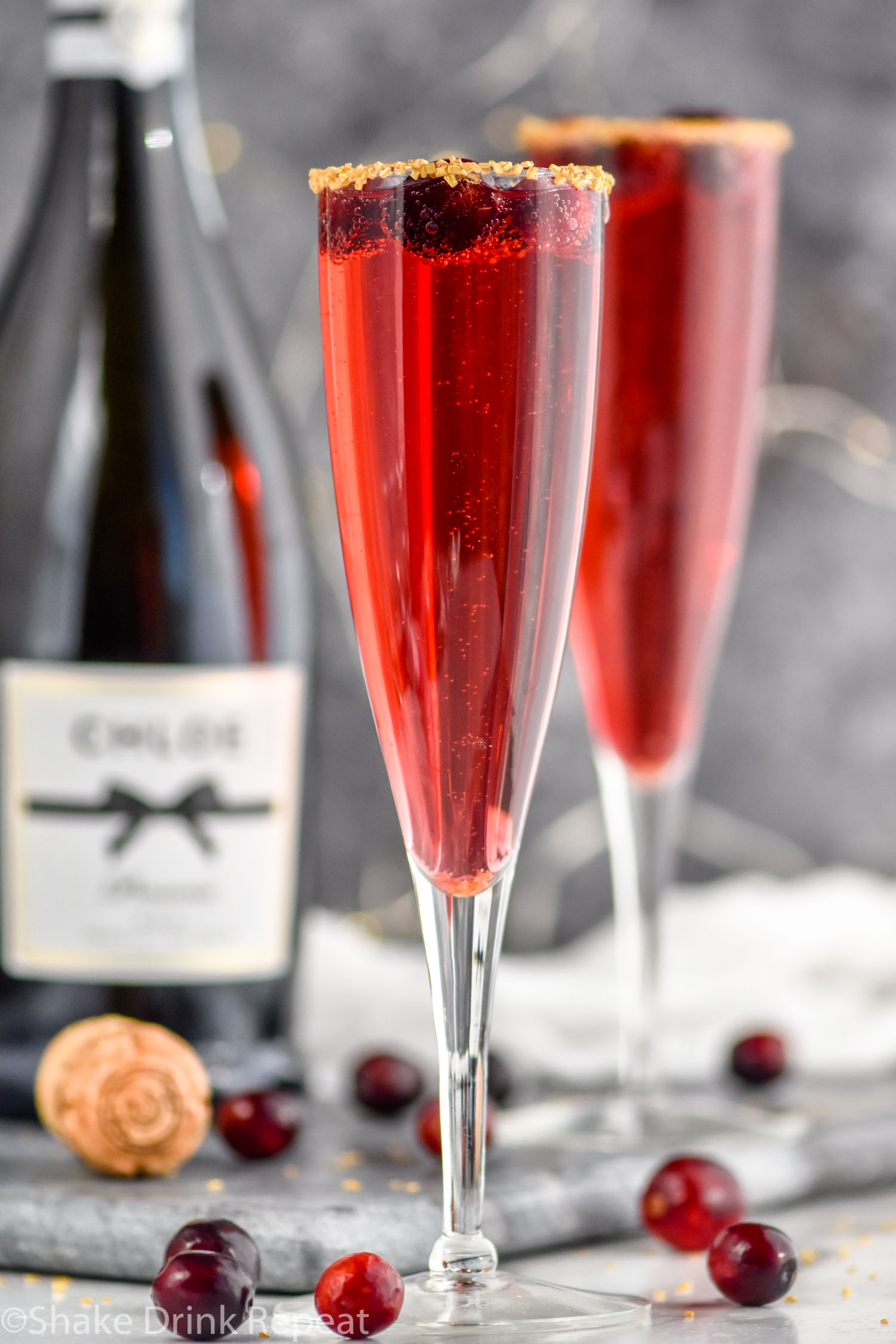 two champagne flutes of Poinsettia Cocktail recipe garnished with gold sprinkles and cranberries with bottle of champagne in the background