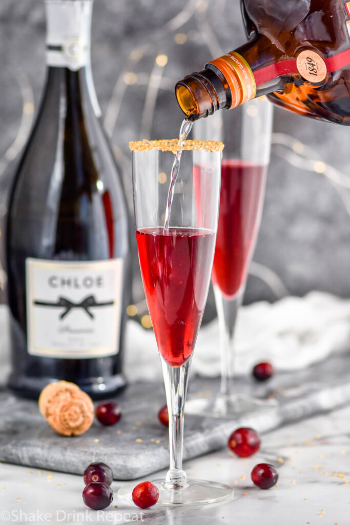 pouring a bottle of Cointreau into a glass of Poinsettia Cocktail recipe garnished with gold sprinkles and cranberries