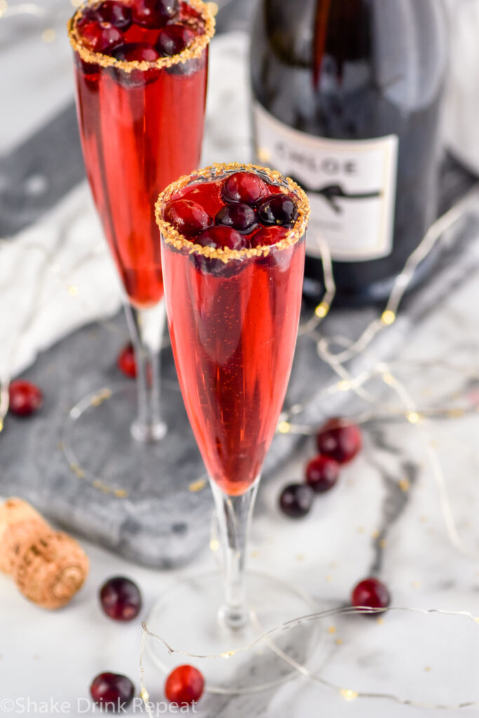 two champagne flutes of Poinsettia Cocktail recipe garnished with gold sprinkles and cranberries with bottle of champagne in the background