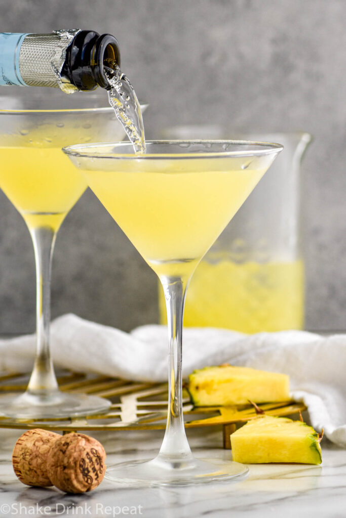 bottle of champagne pouring to a martini glass of Flirtini recipe surrounded by fresh pineapple slices