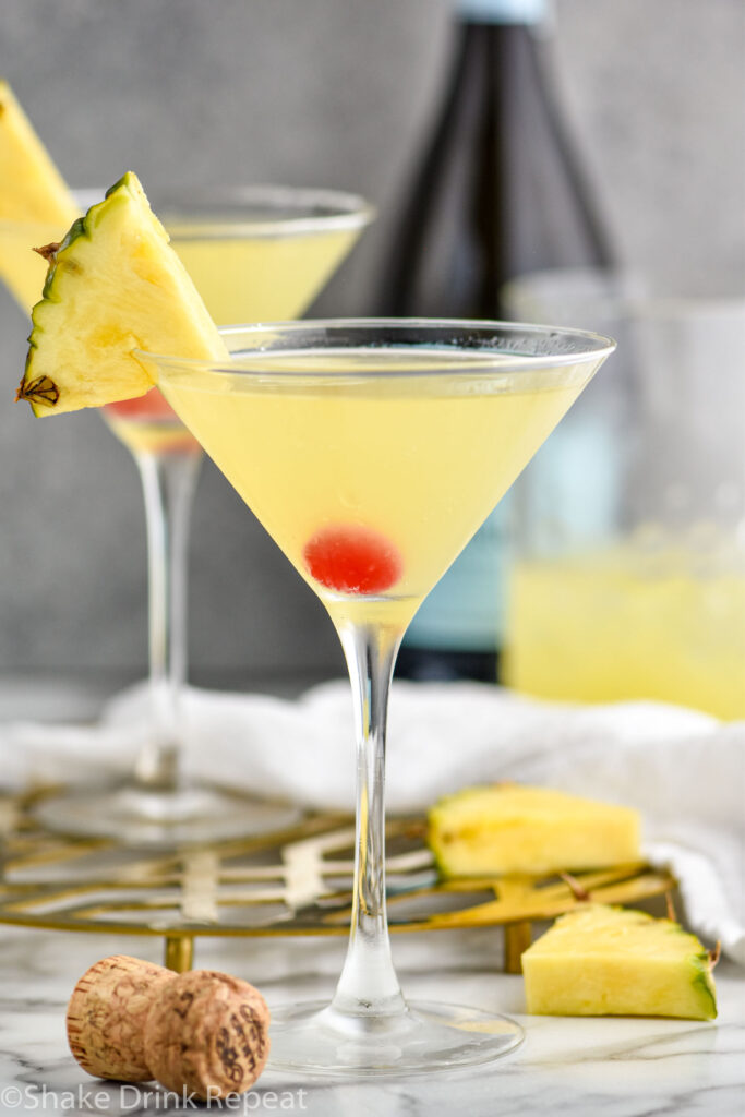 two martini glasses of Flirtini recipe garnished with fresh pineapple and a cherry with bottle of champagne in the background