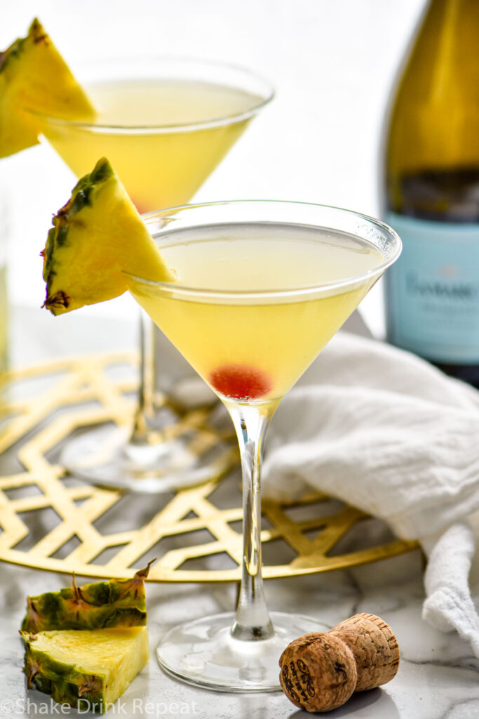 two martini glasses of Flirtini recipe garnished with fresh pineapple slices and bottle of champagne in the background