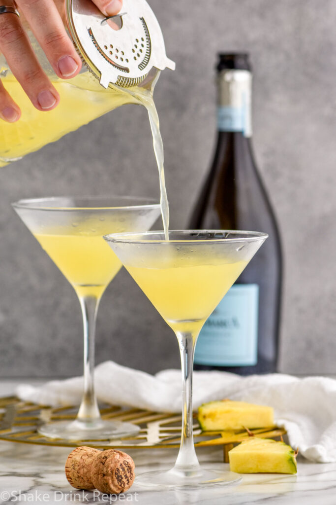 man's hand pouring cocktail mixer of Flirtini ingredients into a chilled martini glass surrounded by bottle of champagne and fresh pineapple slices