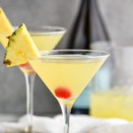 two martini glasses of Flirtini recipe garnished with pineapple slices and a cherry with bottle of champagne in the background
