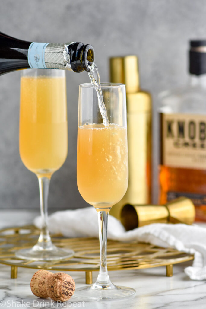 bottle of champagne pouring into a glass of French 95 recipe with bottle of bourbon and cocktail shaker in the background