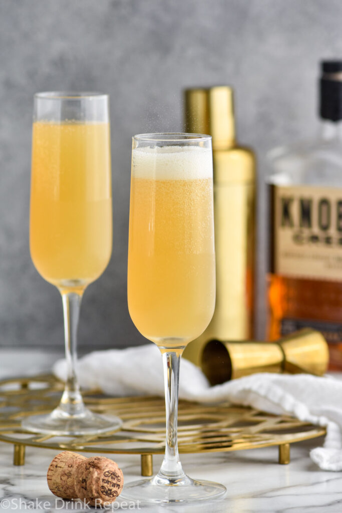 two champagne flutes of French 95 recipe garnished with an orange twist with bottle of bourbon and a cocktail shaker in the background