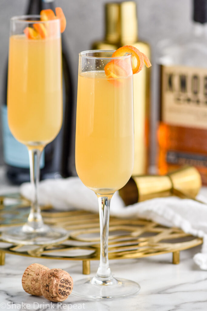 two champagne flutes of French 95 recipe garnished with an orange twist with bottles of bourbon and champagne in the background