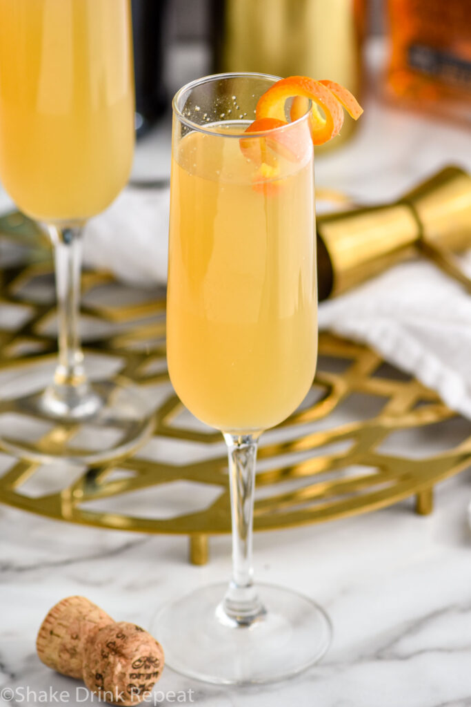 two champagne flutes of French 95 recipe garnished with an orange twist with bottles of bourbon and champagne in the background