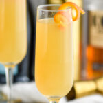 two champagne flutes of French 95 recipe garnished with an orange twist with bottles of bourbon and champagne in the background
