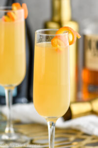 two champagne flutes of French 95 recipe garnished with an orange twist with bottles of bourbon and champagne in the background