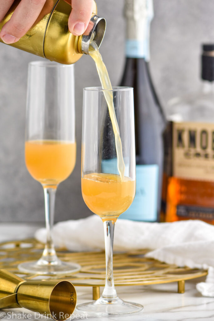 man's hand pouring cocktail shaker of French 95 ingredients into a champagne flute with bottles of champagne and bourbon in the background