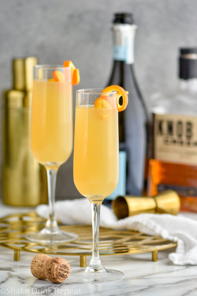 two champagne flutes of French 95 recipe garnished with an orange twist with bottles of bourbon and champagne in the background