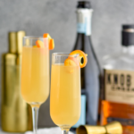 two champagne flutes of French 95 recipe garnished with an orange twist with bottles of bourbon and champagne in the background