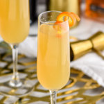 champagne flute of French 95 recipe garnished with an orange twist with bottle of bourbon, cocktail shaker, and jigger to make drink in the background