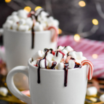 two mugs of boozy peppermint hot chocolate garnished with marshmallows, chocolate drizzle, and candy canes