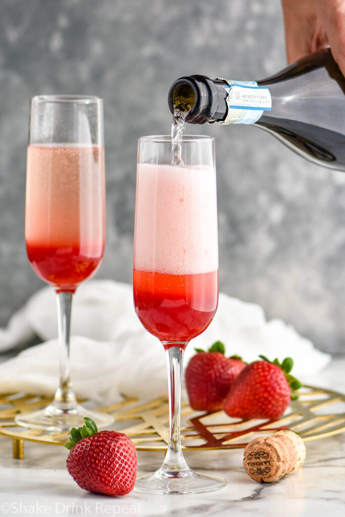 Man's hand pouring bottle of Prosecco into a champagne flute glass of Rossini ingredients surrounded by fresh strawberries