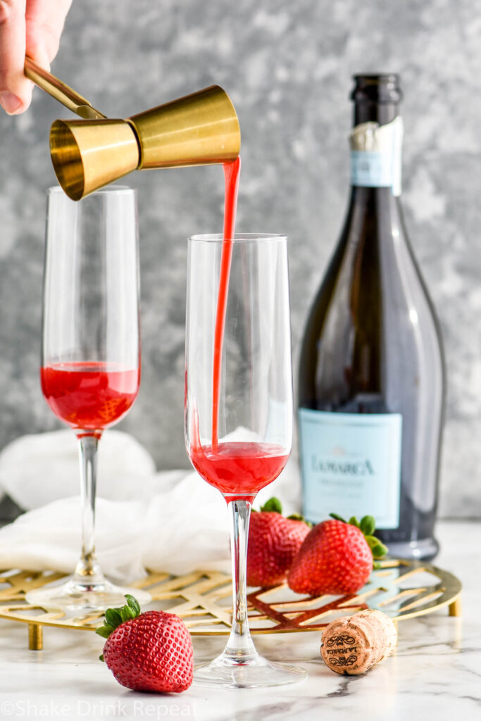 Jigger of strawberry puree pouring into a champagne flute to make a Rossini recipe surrounded by fresh strawberries and a bottle of champagne
