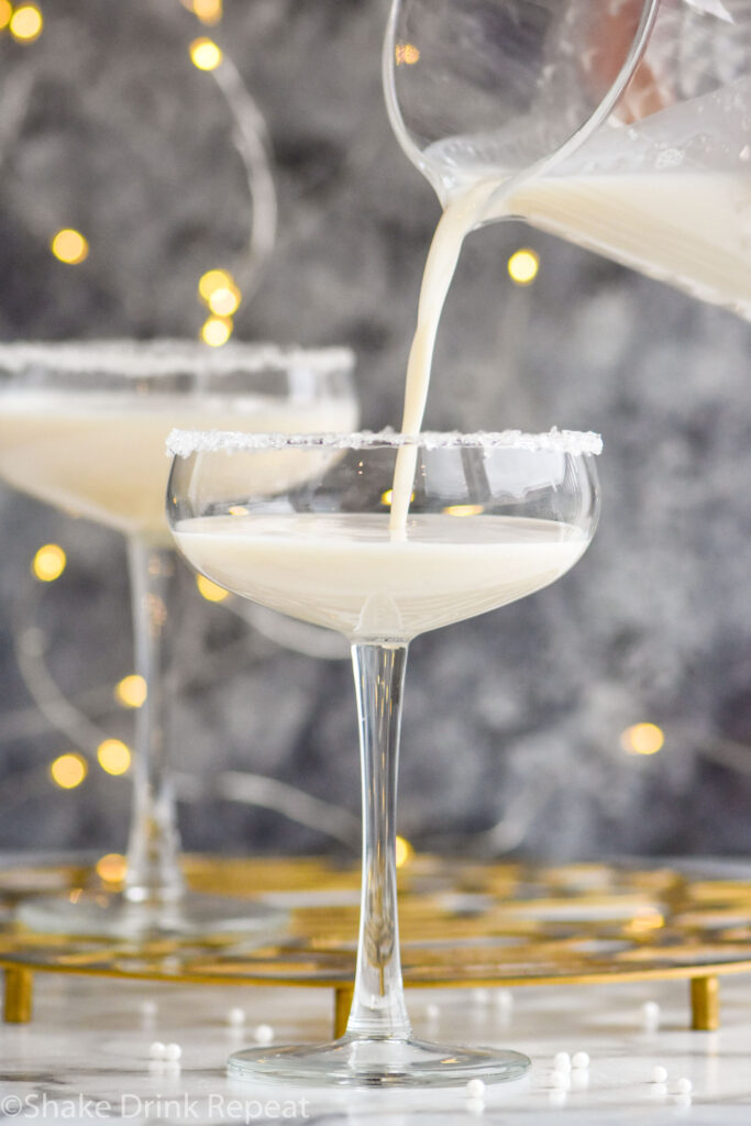 cocktail mixing glass of White Christmas Martini ingredients pouring into coupe glass garnished with white sprinkles