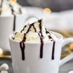 two mugs of Baileys Hot Chocolate garnished with whipped cream and chocolate syrup drizzle surrounded by marshmallows