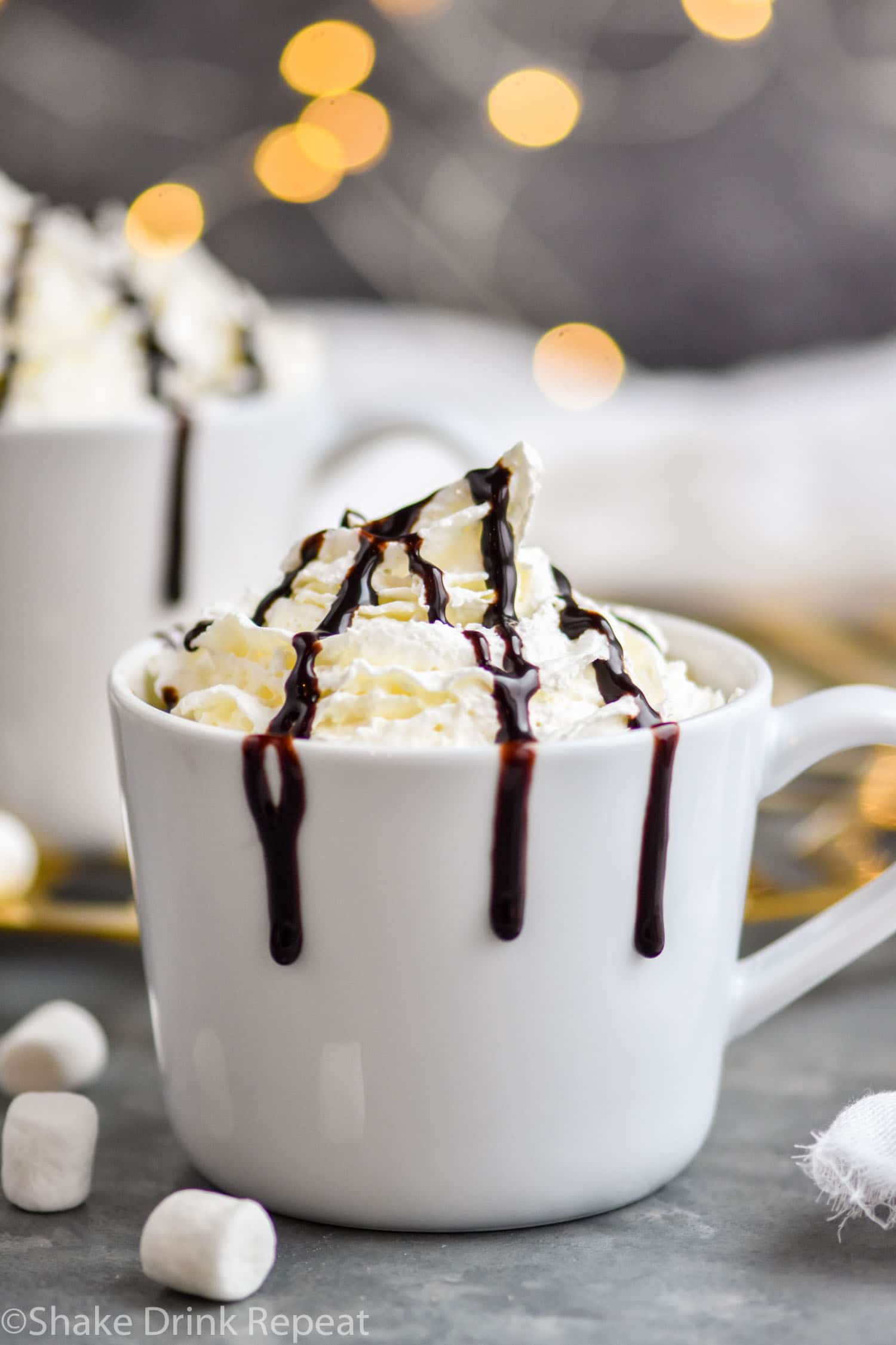 two mugs of Baileys Hot Chocolate garnished with whipped cream and chocolate syrup drizzle surrounded by marshmallows
