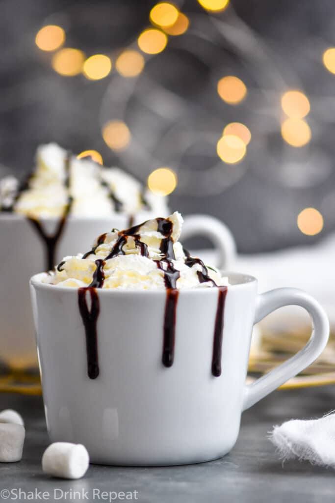 two mugs of Baileys Hot Chocolate garnished with whipped cream and chocolate syrup drizzle surrounded by marshmallows
