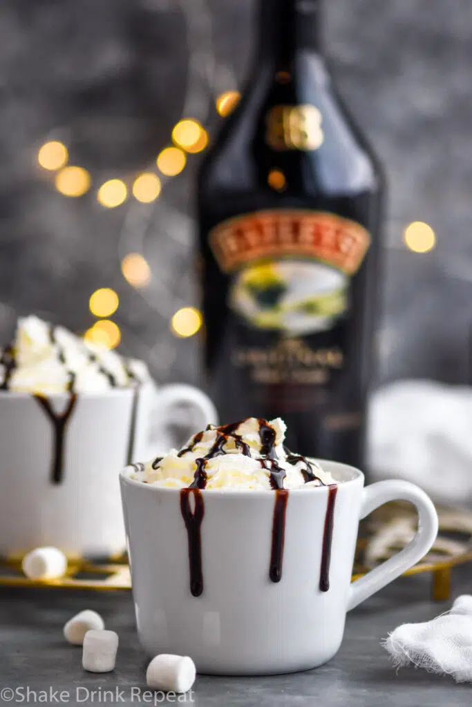 two mugs of Baileys Hot Chocolate garnished with whipped cream and chocolate syrup drizzle surrounded by marshmallows and a bottle of Baileys Irish Cream