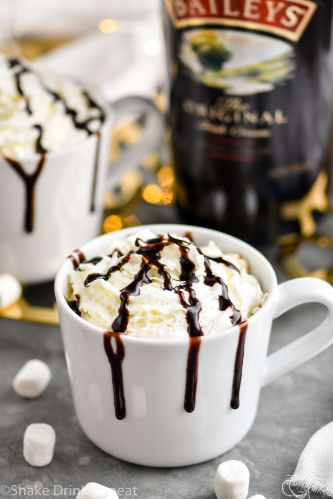 two mugs of Baileys Hot Chocolate garnished with whipped cream and chocolate syrup drizzle surrounded by marshmallows and a bottle of Baileys Irish Cream