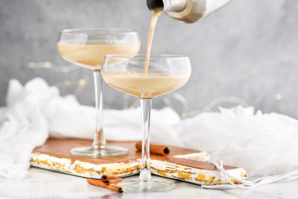 cocktail shaker pouring Cinnamon Roll Martini ingredients into a glass surrounded by cinnamon sticks