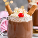 two mugs of Drunk Reindeer recipe garnished with whipped cream, cinnamon sticks, a cherry, and chocolate shavings