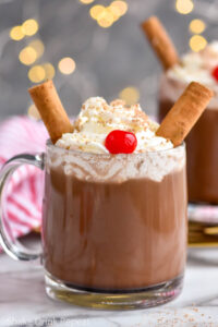 two mugs of Drunk Reindeer recipe garnished with whipped cream, cinnamon sticks, a cherry, and chocolate shavings