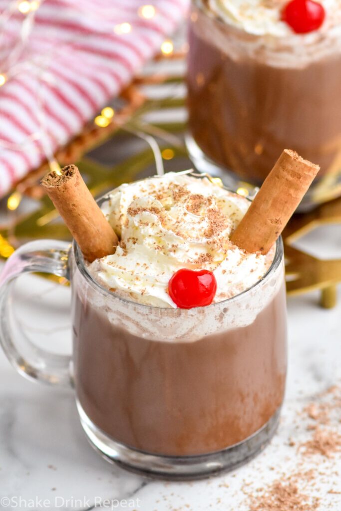 two mugs of Drunk Reindeer recipe garnished with whipped cream, cinnamon sticks, a cherry, and chocolate shavings