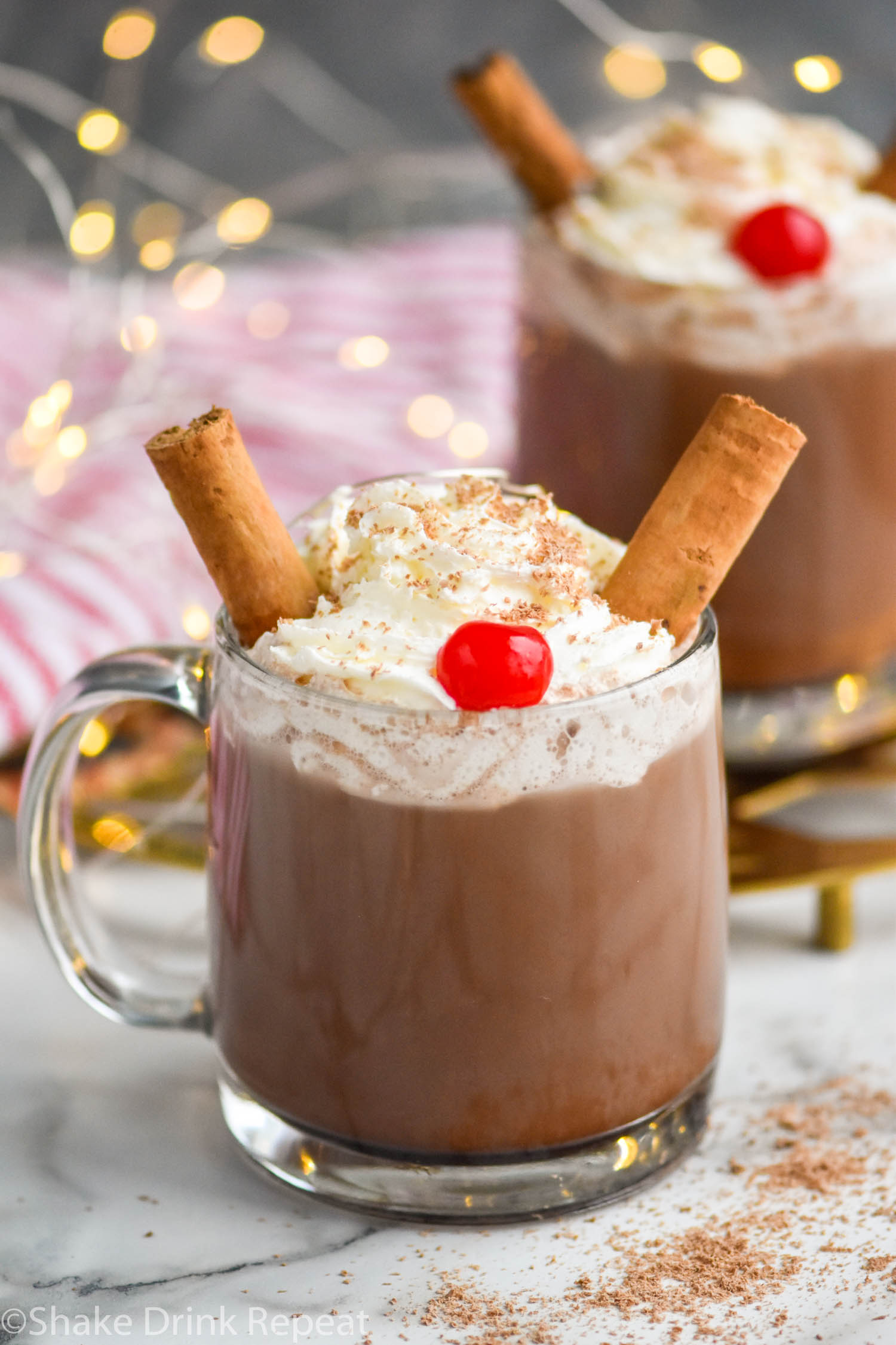 two mugs of Drunk Reindeer recipe garnished with whipped cream, cinnamon sticks, a cherry, and chocolate shavings