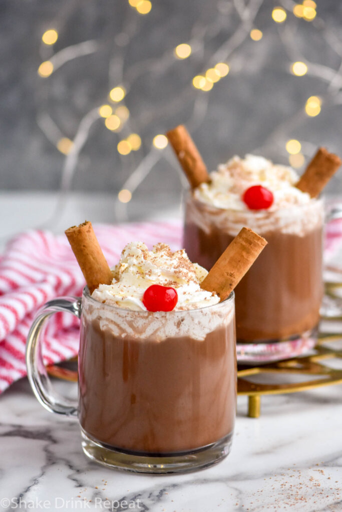 two mugs of Drunk Reindeer recipe garnished with whipped cream, cinnamon sticks, a cherry, and chocolate shavings