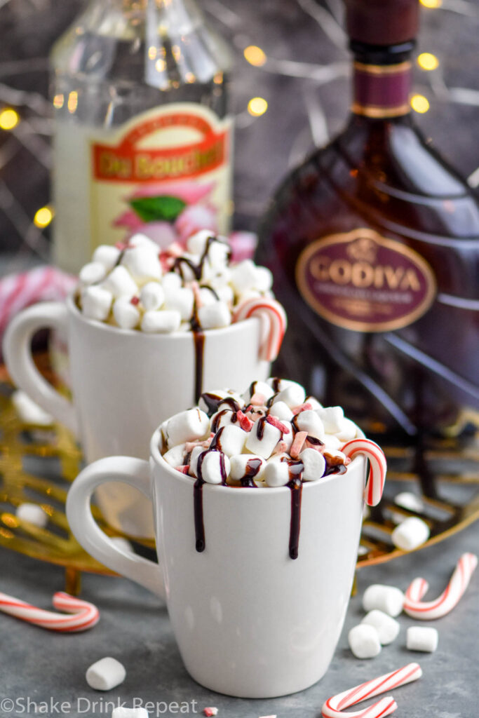 two mugs of boozy peppermint hot chocolate garnished with marshmallows, chocolate drizzle, and candy canes with bottles of peppermint schnapps and Godiva chocolate liqueur in the background
