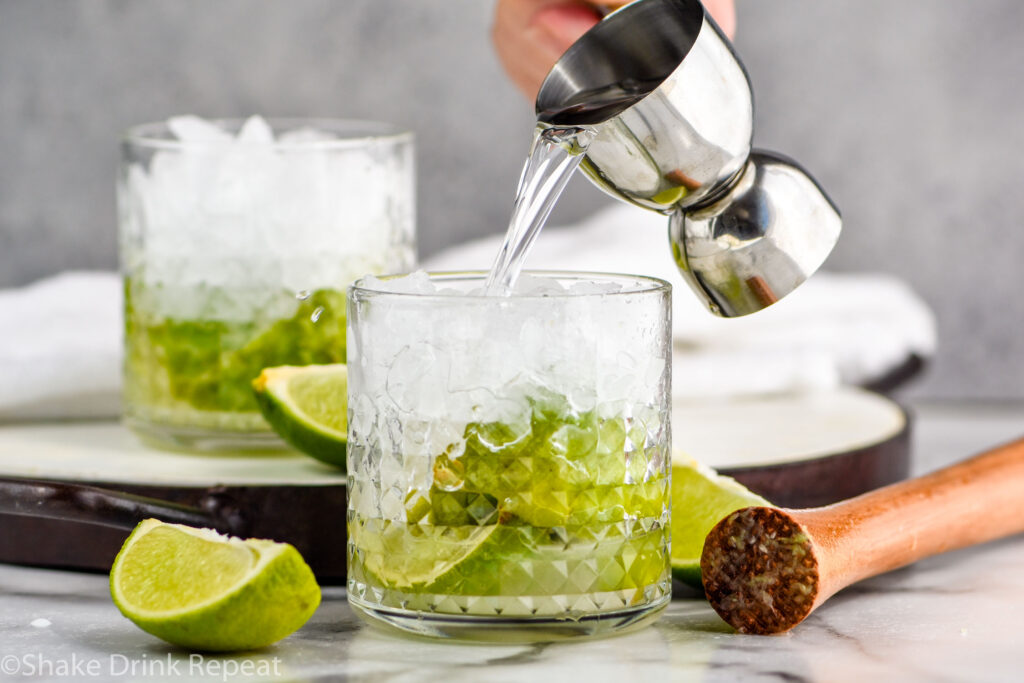 man's hand pouring jigger of vodka into a glass of muddled limes and sugar to make a Caipiroska recipe surrounded by lime wedges
