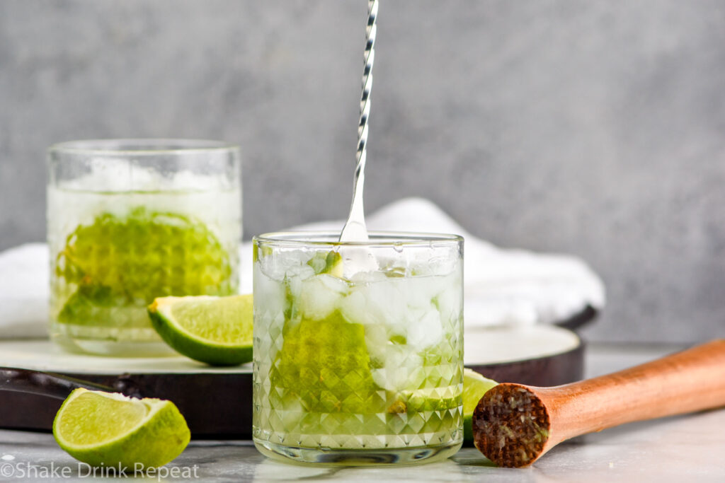 stir stick stirring glass of Caipiroska ingredients with ice surrounded by lime wedges and a cocktail muddler