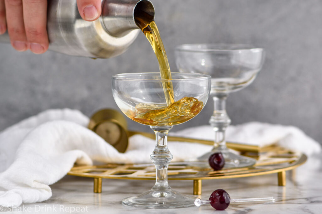 cocktail shaker pouring Corpse Reviver ingredients into a coupe glass surrounded by cocktail spear with cherry