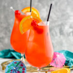 two glasses of Hurricane cocktail recipe with ice garnished with orange slice, cherries, and a straw surrounded by fruit and a drink umbrella