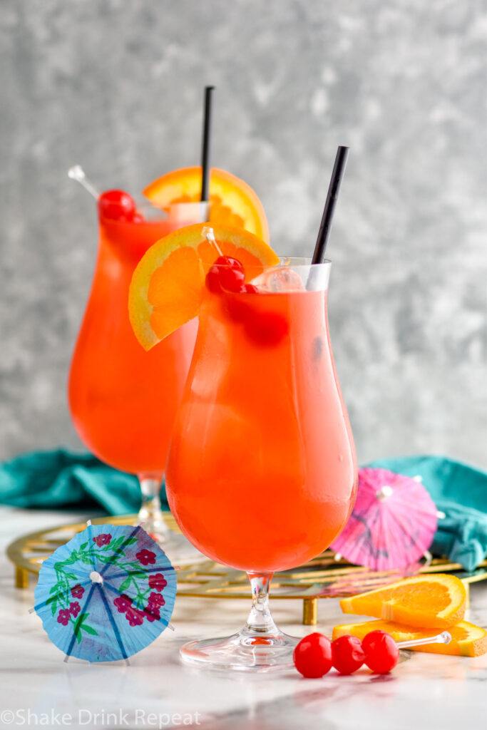 two glasses of Hurricane cocktail recipe with ice garnished with orange slice, cherries, and a straw surrounded by fruit and a drink umbrella