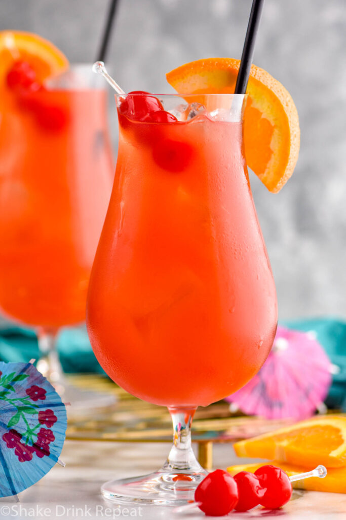 two glasses of Hurricane cocktail recipe with ice garnished with orange slice, cherries, and a straw surrounded by fruit and a drink umbrella