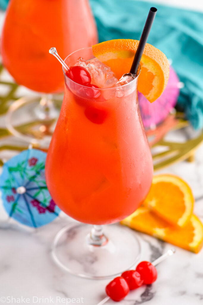 two glasses of Hurricane cocktail recipe with ice garnished with orange slice, cherries, and a straw surrounded by fruit and a drink umbrella