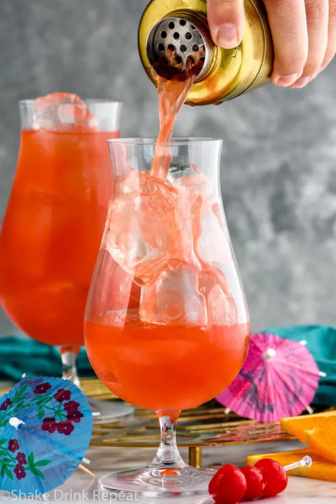 man's hand pouring cocktail shaker of Hurricane ingredients into a glass of ice to make Hurricane recipe garnished with orange slice and cherries