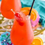 two glasses of Hurricane cocktail recipe with ice garnished with orange slice, cherries, and a straw surrounded by fruit and a drink umbrella